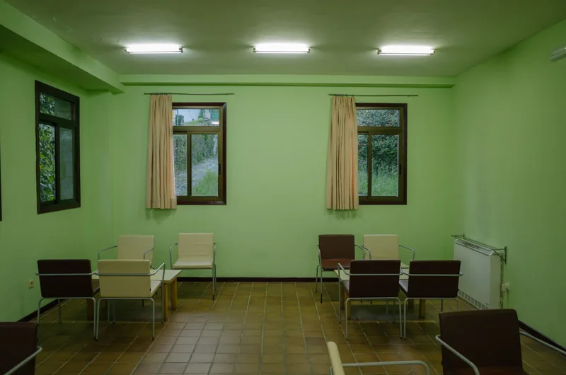 Sala vacía de paredes verdes, con varias sillas dispersas y tres ventanas.