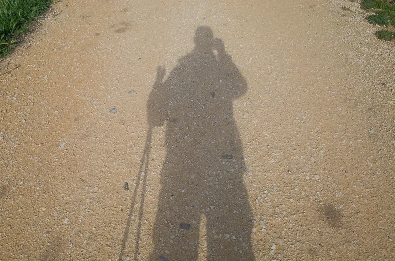 La sombra alargada de una persona con bastones de caminata se proyecta sobre un camino de grava.