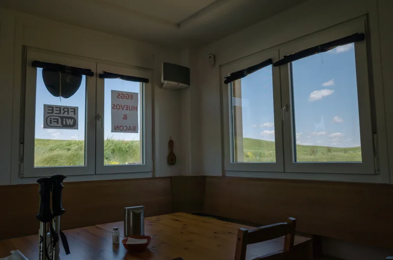 Interior en penumbra con mesas y sillas frente a ventanas que dan a un paisaje verde.