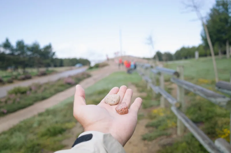 Una mano sostiene dos piedras; al fondo, desenfocados, un sendero y un grupo de caminantes.