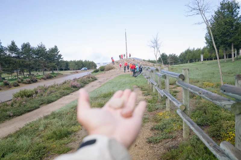 Un grupo de caminantes avanza hacia un monumento; en primer plano, una mano desenfocada sostiene dos piedras.