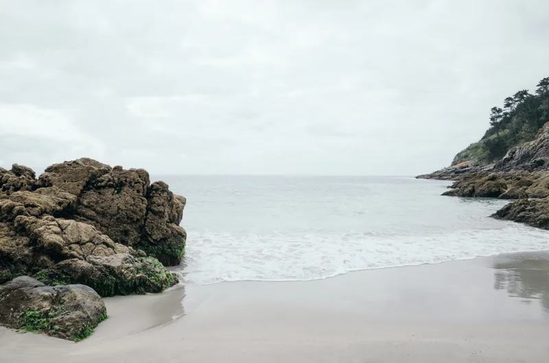 Pequeña cala de arena entre rocas oscuras, frente a un mar calmo y un cielo nublado.